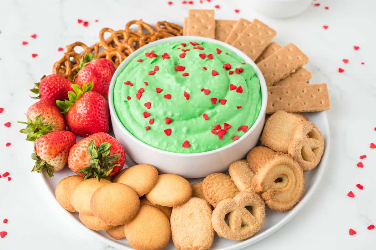 A white plate with strawberries, pretzels, cookies, and graham crackers arranged around a bowl of green dip topped with small red heart sprinkles.