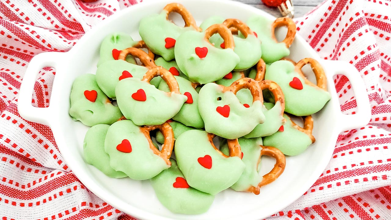 A plate of mini pretzels coated in green chocolate, each topped with a small red heart-shaped sprinkle, sits on a red and white striped cloth.