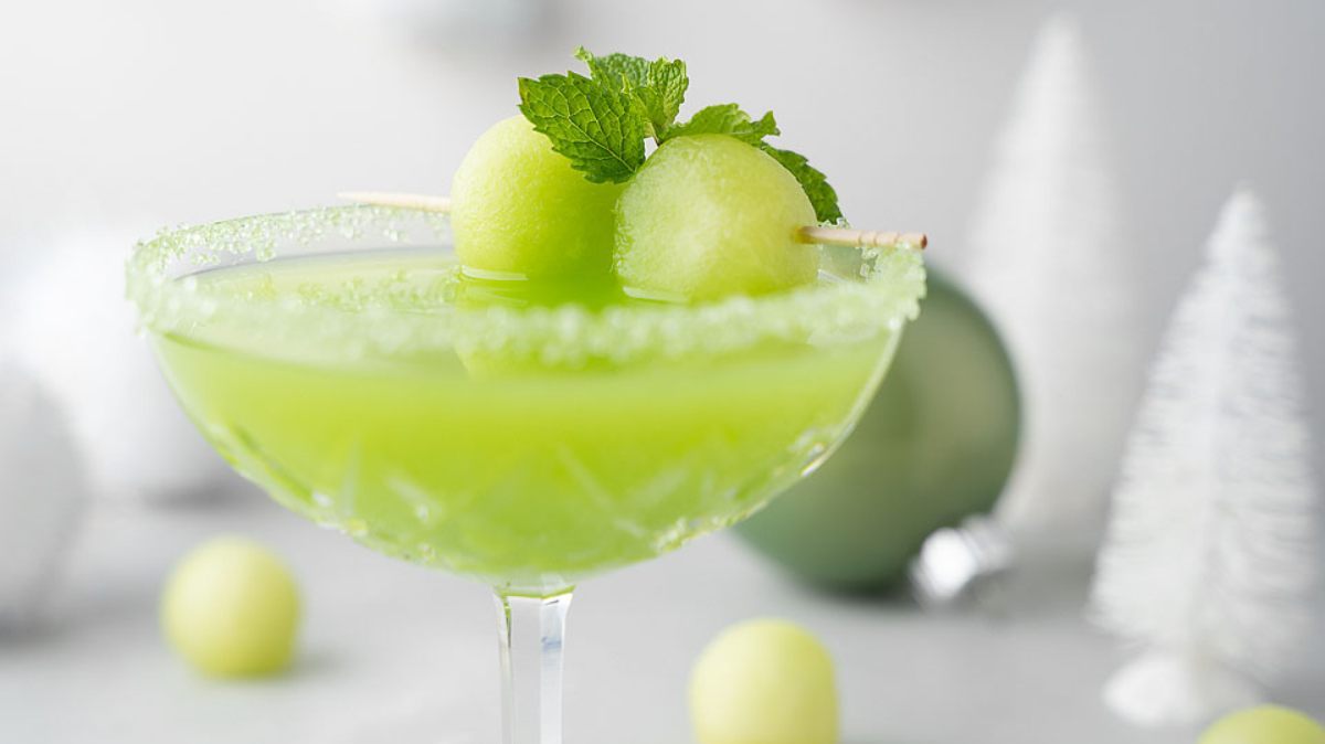 A green cocktail in a glass with a sugared rim, garnished with two melon balls and a sprig of mint on a skewer. White decorative trees and ornaments are in the blurred background.