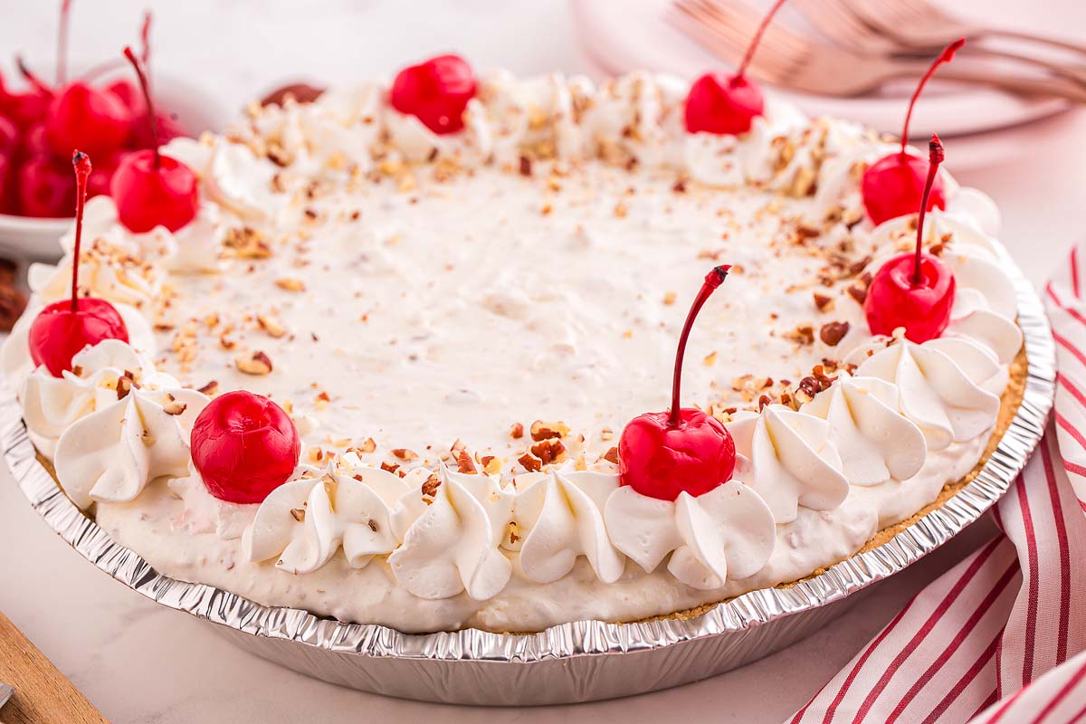 A cream pie in a foil pan topped with whipped cream swirls, maraschino cherries, and chopped nuts, with a red and white striped napkin nearby.