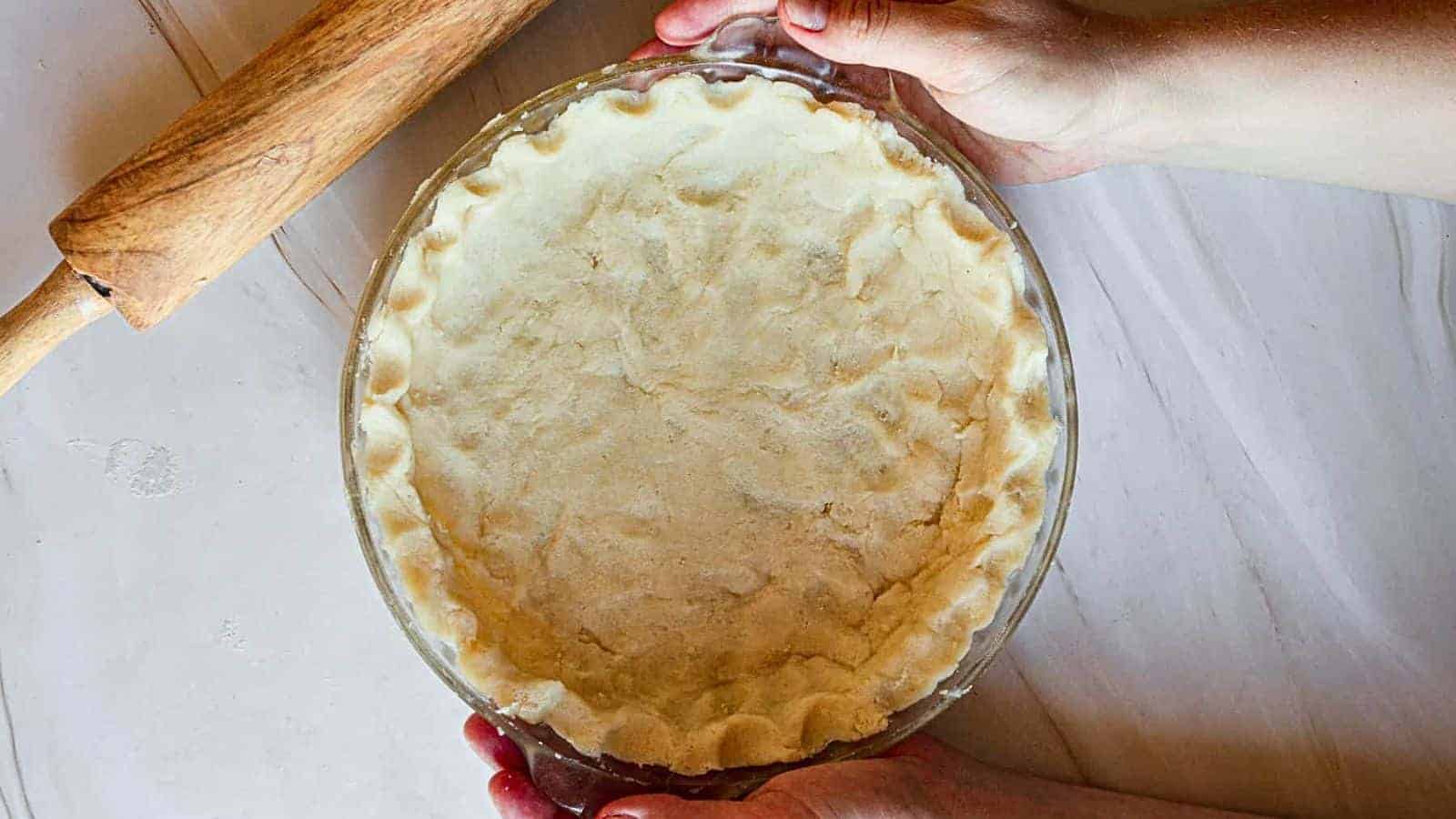 Hands holding a glass pie dish with an unbaked pie crust next to a wooden rolling pin on a light-colored surface.