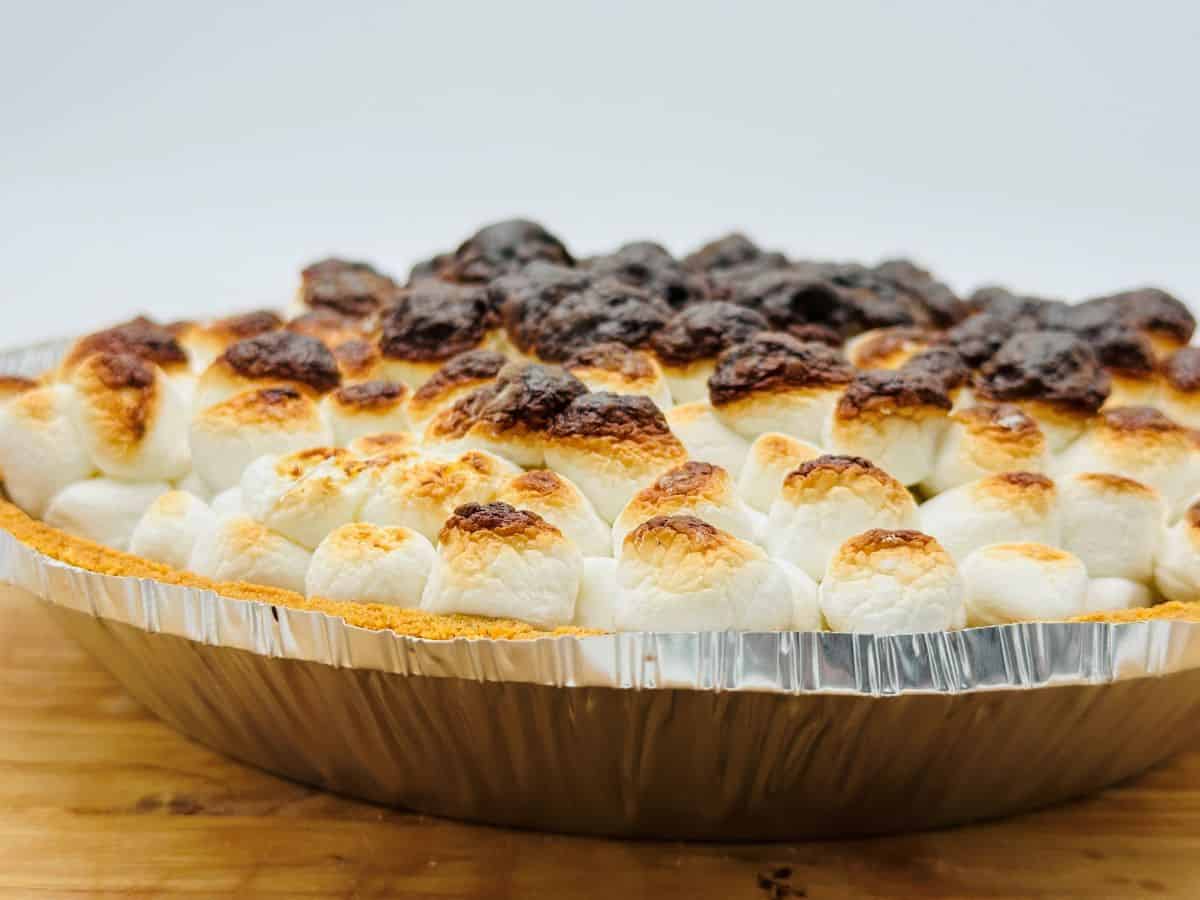 A close-up of a pie in a foil tin topped with toasted marshmallows, some of which are browned and slightly burnt.