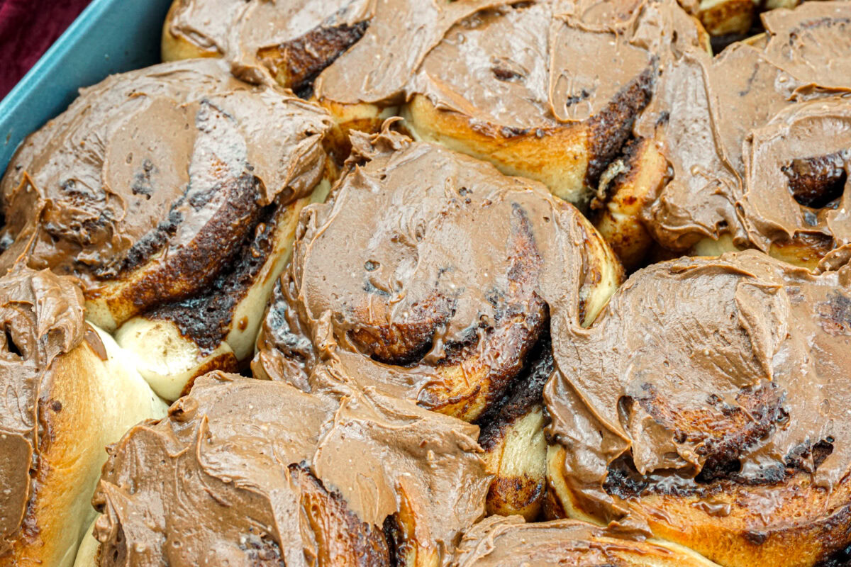 A close-up view of freshly baked cinnamon rolls in a pan, topped with a layer of chocolate frosting.