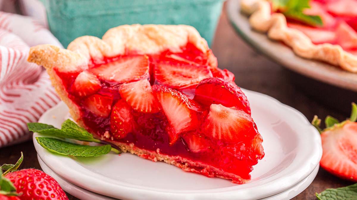 A slice of strawberry pie with a flaky crust and fresh strawberry topping sits on a white plate, garnished with mint leaves.