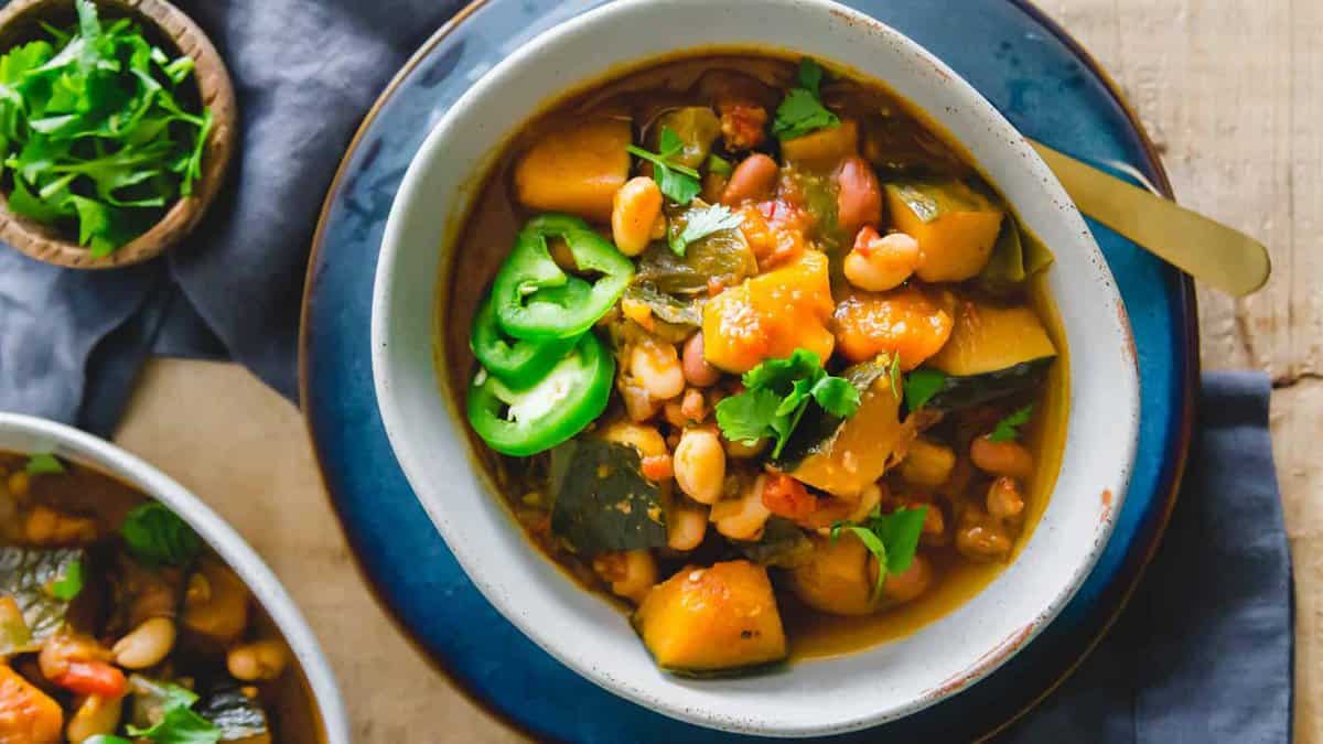 A bowl of vegetable stew with beans, squash, greens, and sliced jalapeรฑos, garnished with fresh cilantro, is placed on a blue plate next to a small bowl of cilantro.