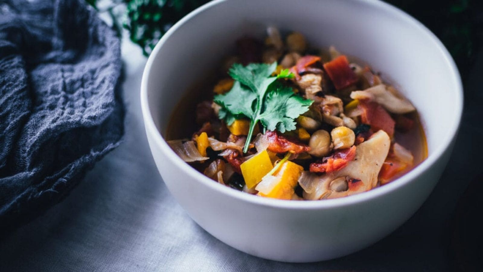 A white bowl filled with vegetable and chickpea stew, garnished with fresh cilantro, sits on a table next to a dark cloth.