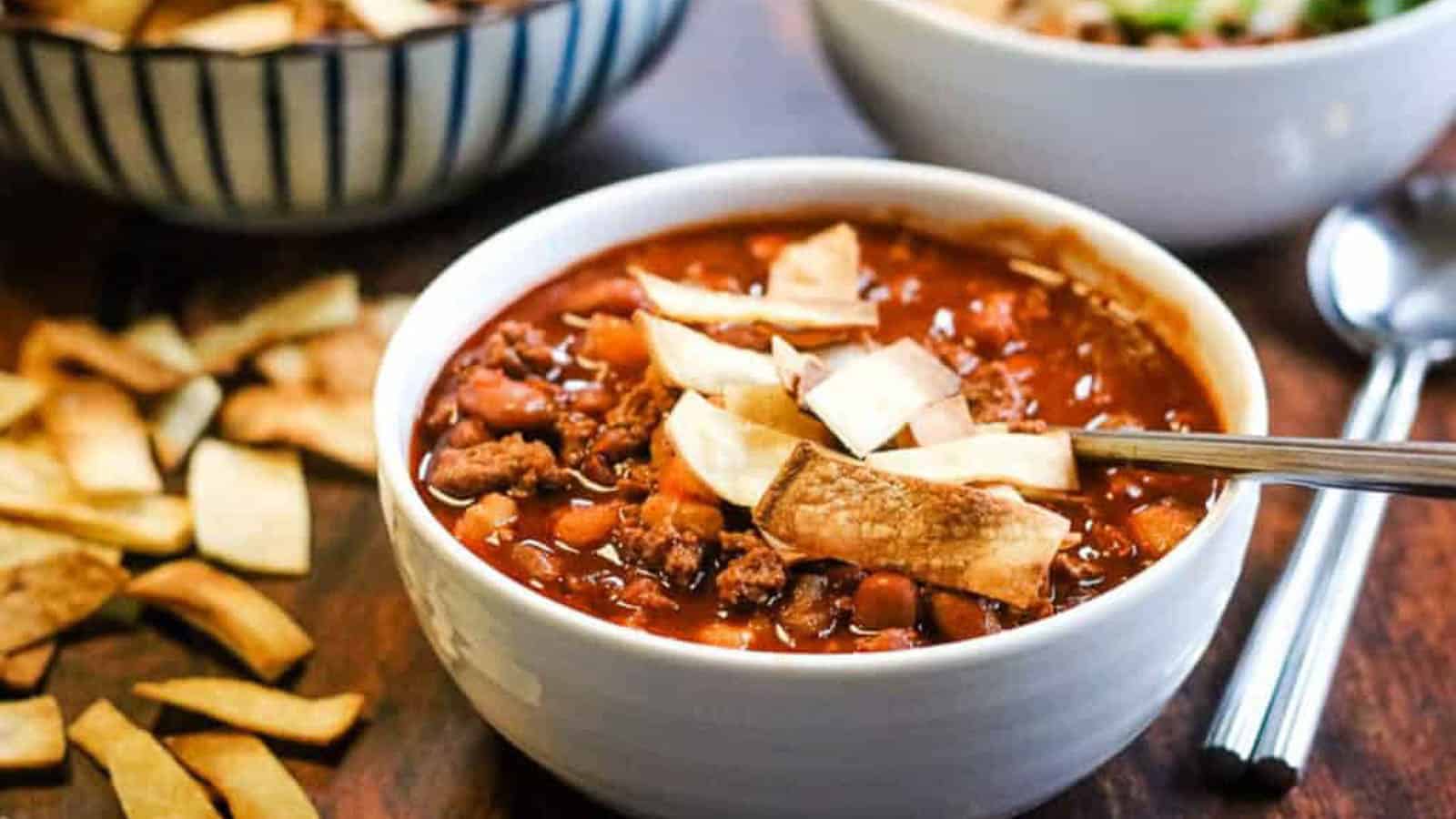 A bowl of chili topped with tortilla strips sits on a wooden surface next to a spoon, with another bowl and extra tortilla strips in the background.
