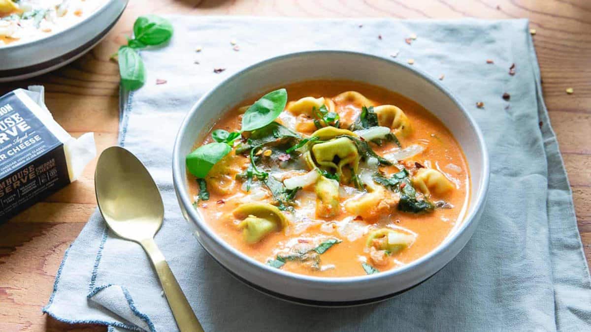 A bowl of creamy tomato soup with tortellini pasta and fresh basil, placed on a gray cloth with a gold spoon on the side.