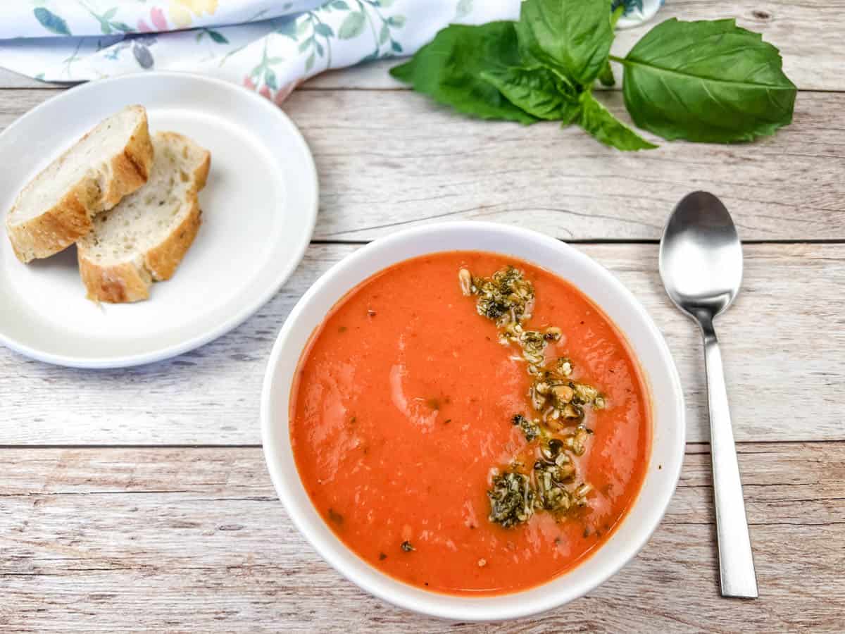 A bowl of tomato soup with herbs on top, next to a spoon and a plate with two slices of bread on a wooden surface. Fresh basil leaves are in the background.