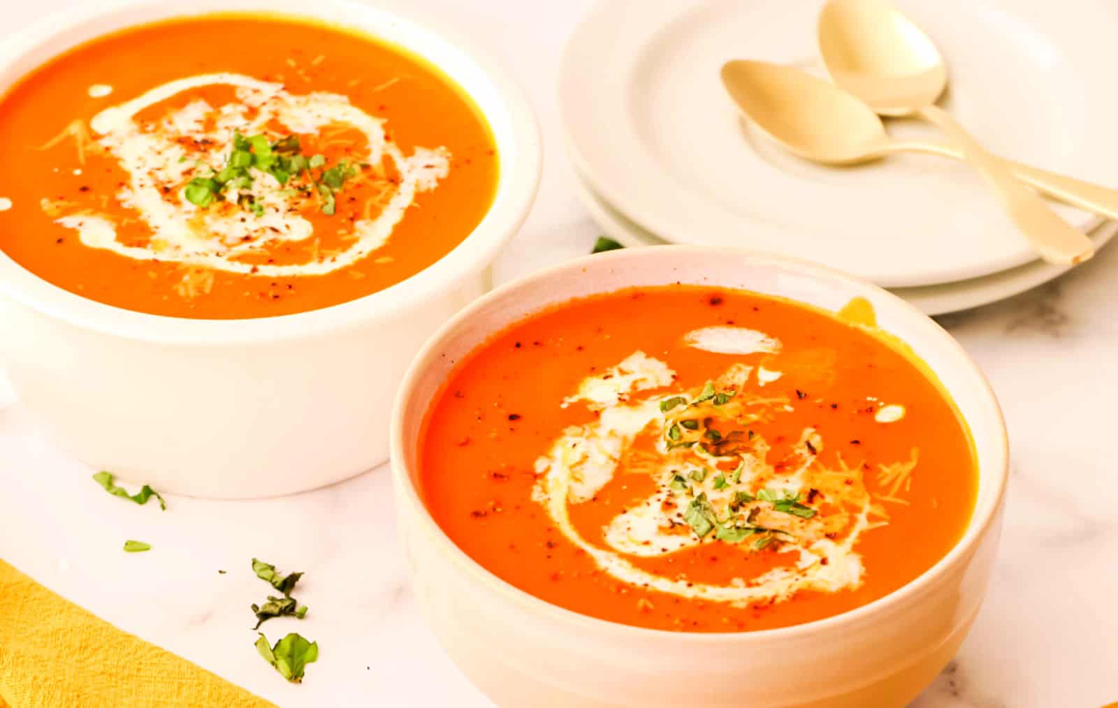 Two bowls of creamy tomato soup garnished with a swirl of cream and chopped herbs, placed on a white surface next to stacked plates and spoons.