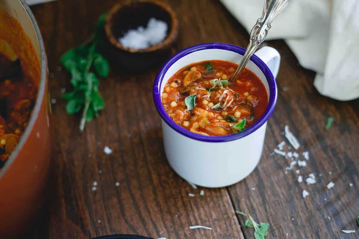 A white enamel mug filled with tomato-based bean soup sits on a wooden table, with a spoon inside and fresh herbs sprinkled on top.