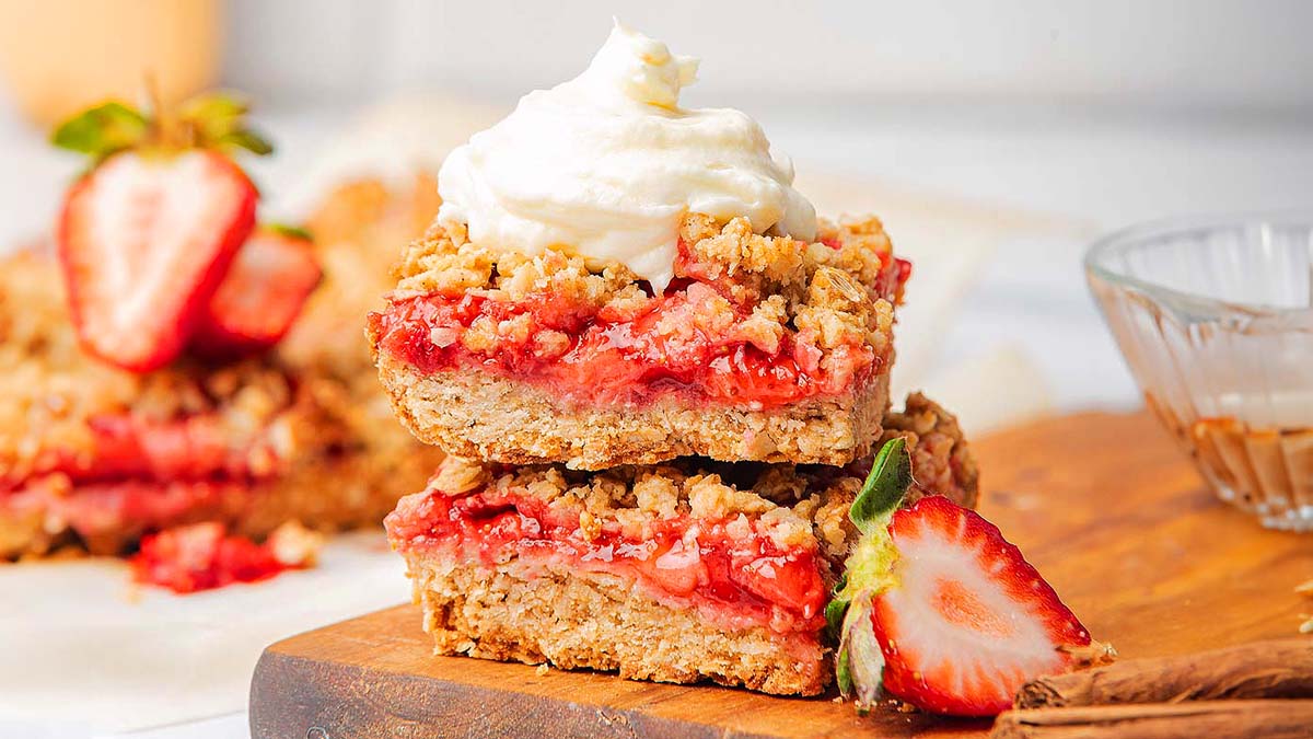 Two stacked strawberry crumble bars topped with a dollop of whipped cream, garnished with fresh strawberry halves on a wooden board.