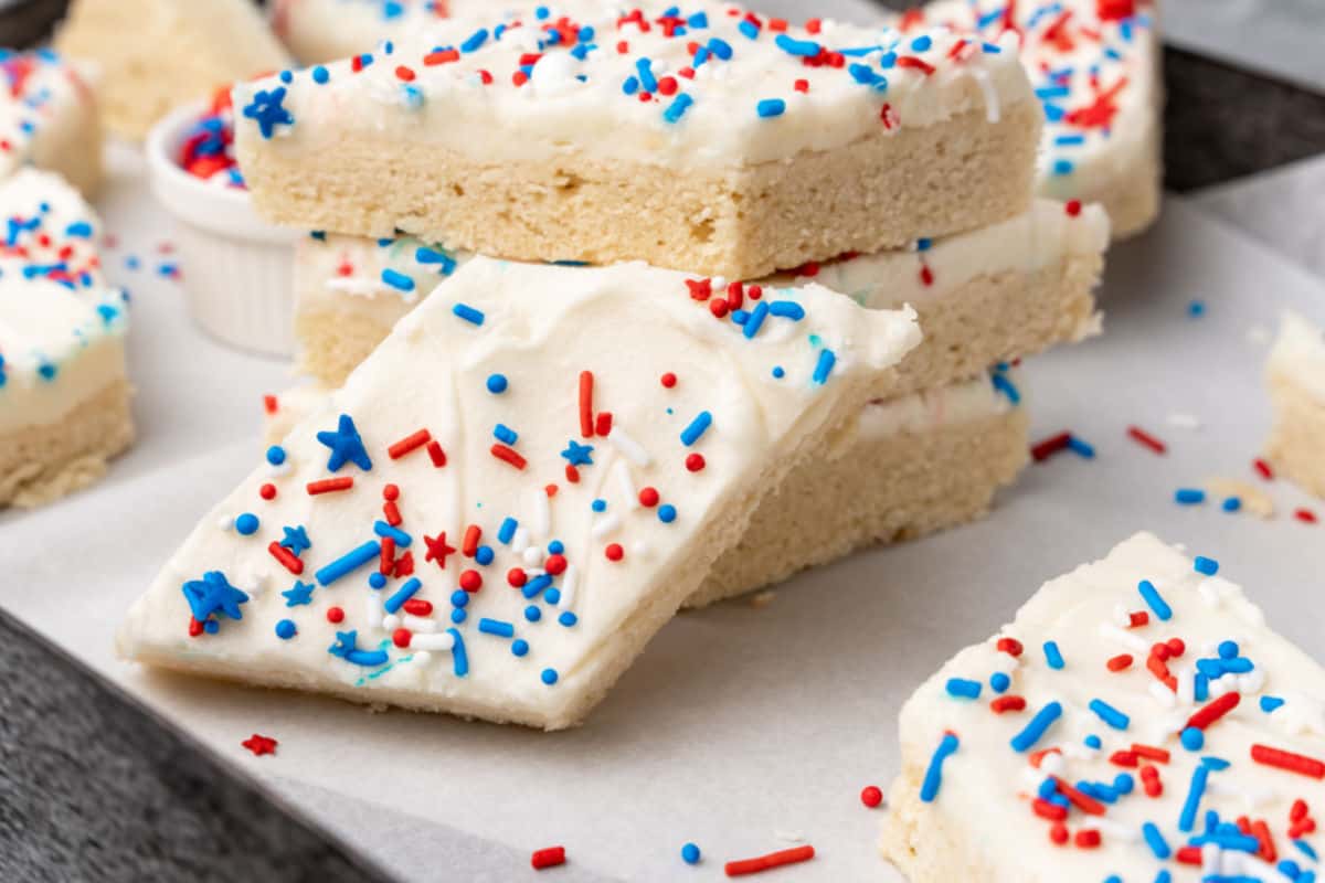 Frosted sugar cookie bars with white icing and red, white, and blue sprinkles are stacked on parchment paper.