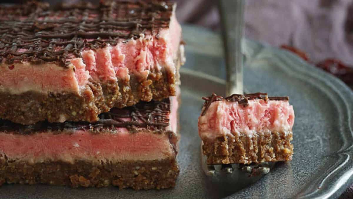A fork holds a bite-sized piece of a layered dessert bar with a crumbly brown base, pink middle layer, and chocolate drizzle on top, served on a gray plate.