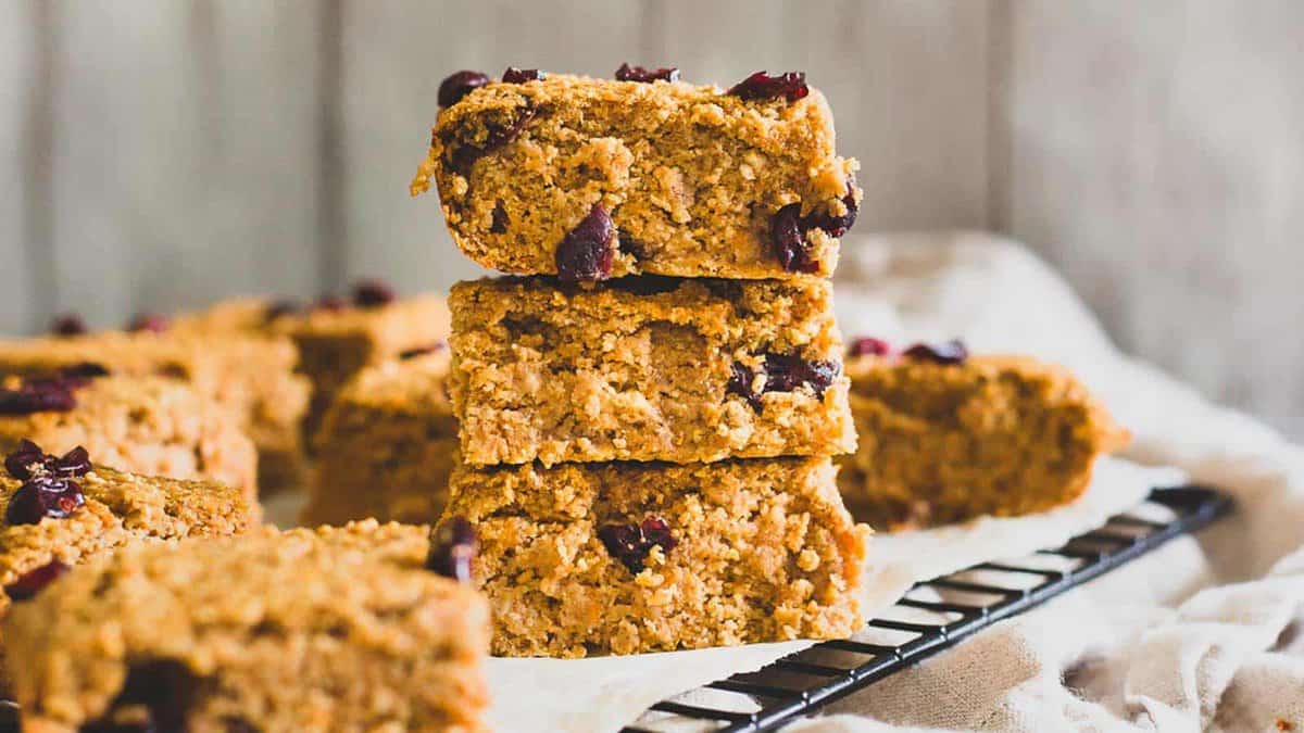 Three oat bars with visible dried cranberries are stacked on a cooling rack, surrounded by more bars, against a neutral background.