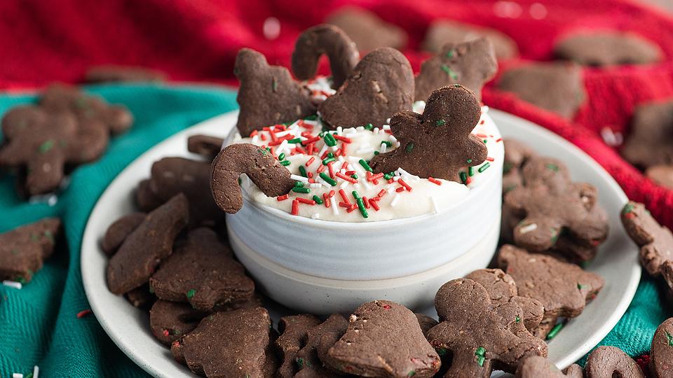 A bowl of white dip with red and green sprinkles is surrounded by chocolate holiday-shaped cookies on a plate, with red and green fabric in the background.