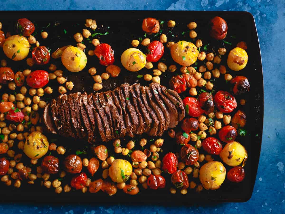Sliced roast beef on a black tray, surrounded by roasted cherry tomatoes, yellow potatoes, and chickpeas.