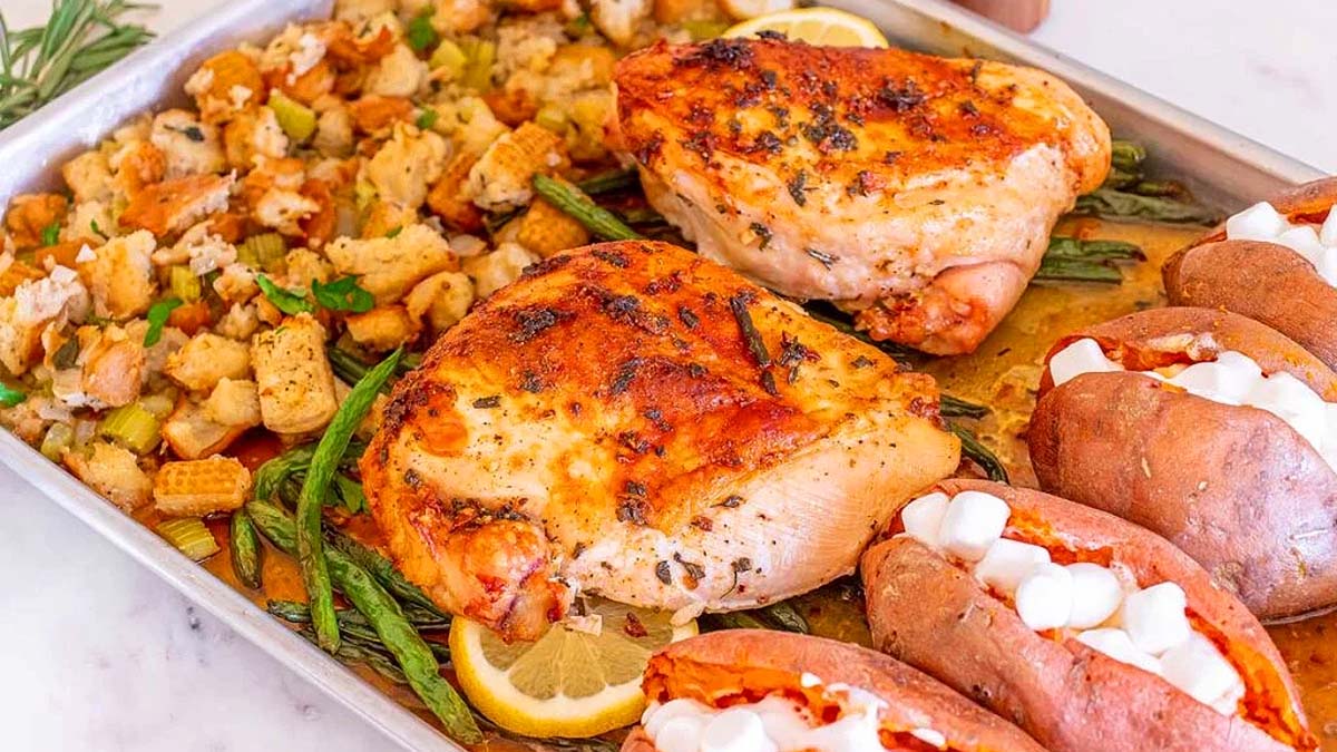 A sheet pan with roasted chicken breasts on green beans, baked sweet potatoes topped with marshmallows, and a serving of stuffing with herbs.