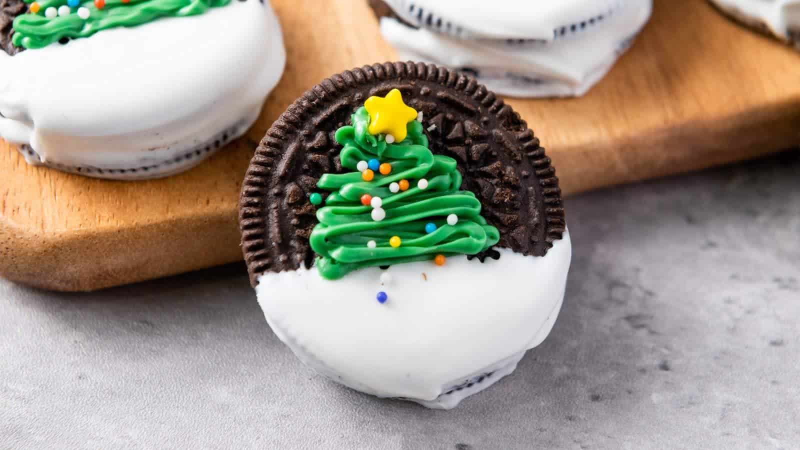 A chocolate sandwich cookie half-dipped in white icing, decorated with green frosting shaped like a Christmas tree and topped with colorful sprinkles and a yellow star.