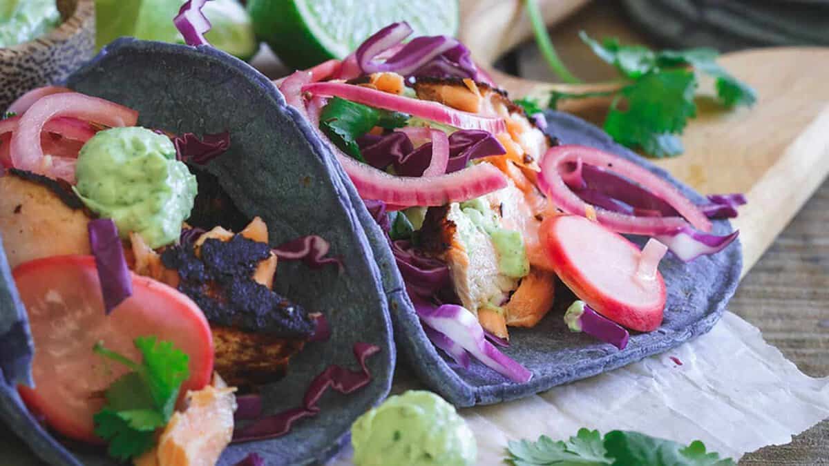 Two blue corn tacos filled with grilled chicken, sliced radishes, red cabbage, pickled onions, cilantro, and green sauce are displayed on parchment paper.