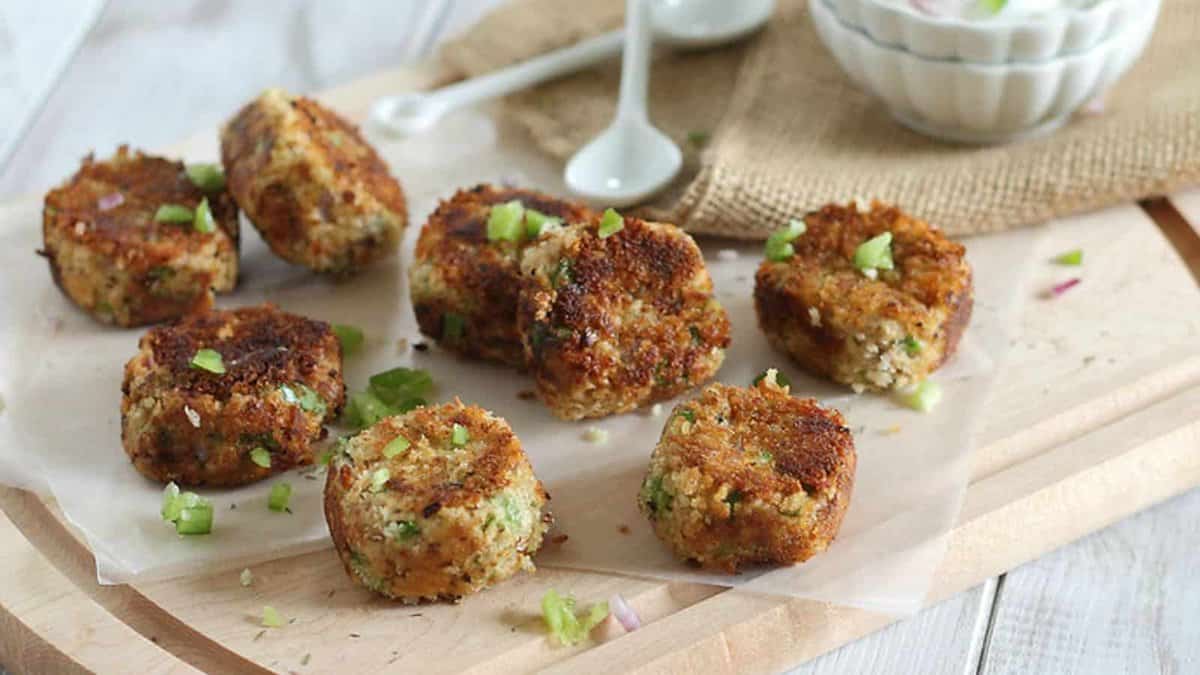 Eight golden-brown vegetable cutlets garnished with chopped green herbs are arranged on parchment paper atop a wooden board, with a small bowl and spoons in the background.