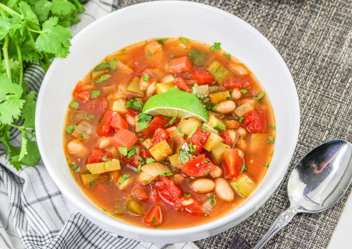 A bowl of vegetable and bean soup garnished with a lime wedge, placed on a white plate next to a spoon, a checkered napkin, and fresh cilantro.