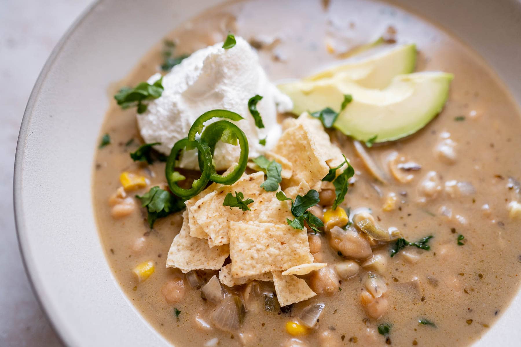 A bowl of creamy soup with white beans, corn, tortilla chips, sliced avocado, jalapeรฑo, cilantro, and a dollop of sour cream.