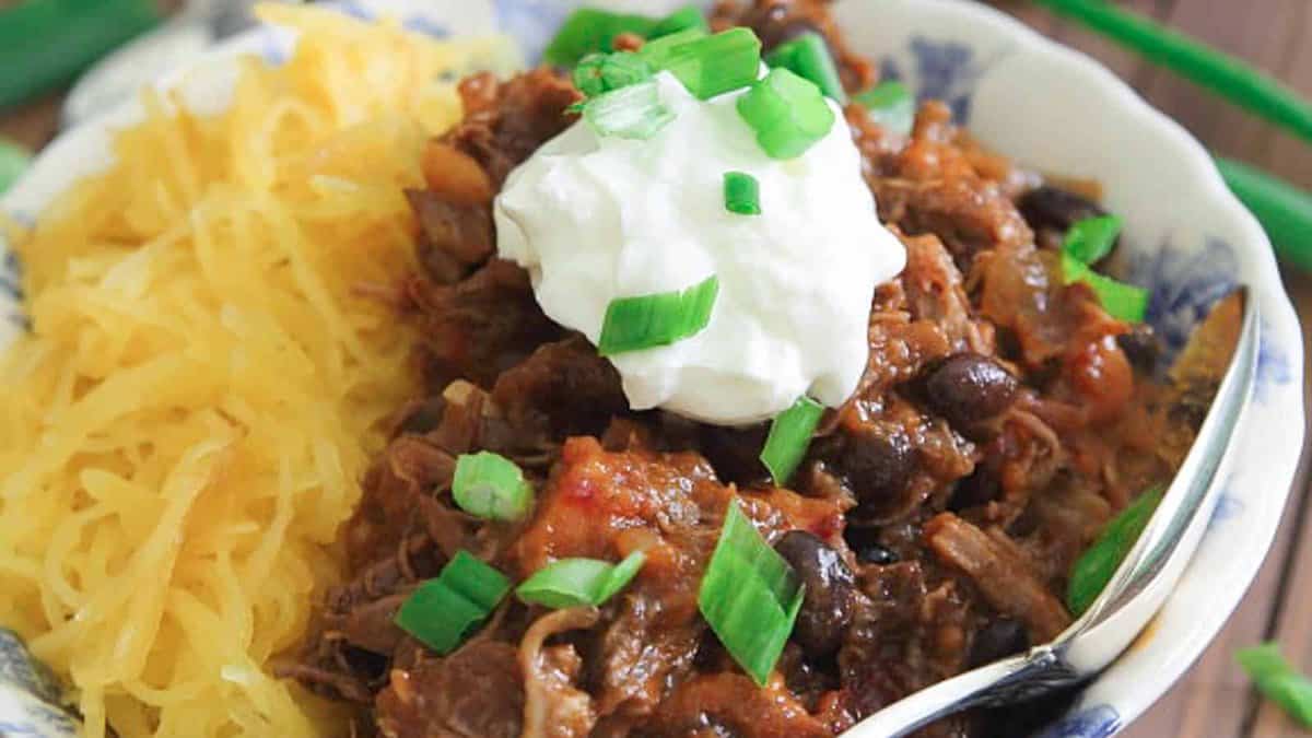 A bowl of spaghetti squash topped with chili, black beans, a dollop of sour cream, and chopped green onions, with a spoon on the side.
