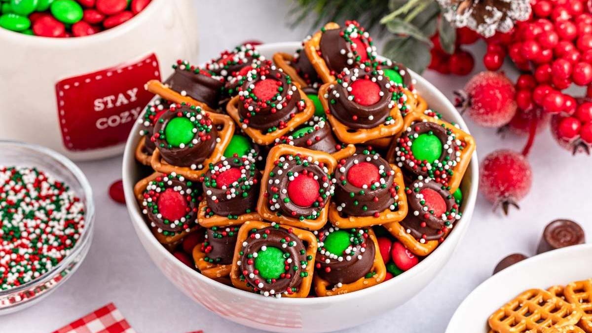 A bowl filled with holiday-themed pretzel bites topped with chocolate, red and green candies, and colorful sprinkles, surrounded by festive decorations and ingredients.