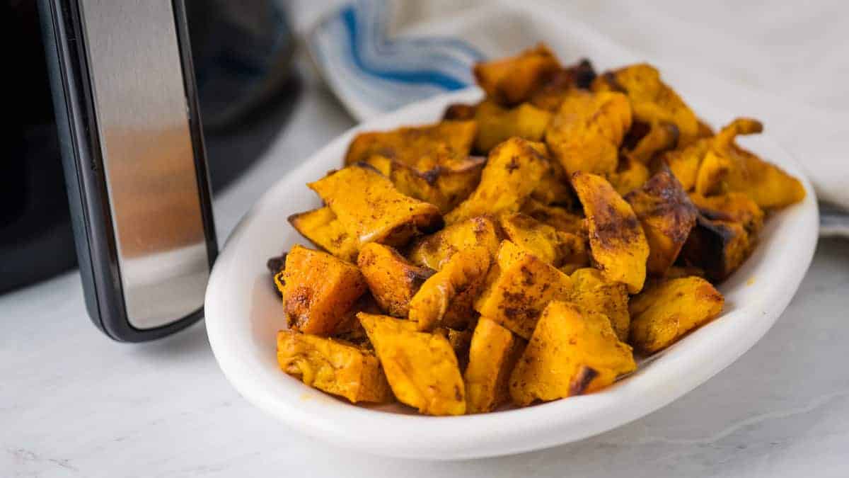 A white oval plate filled with cooked, seasoned sweet potato cubes sits on a marble countertop next to an air fryer.