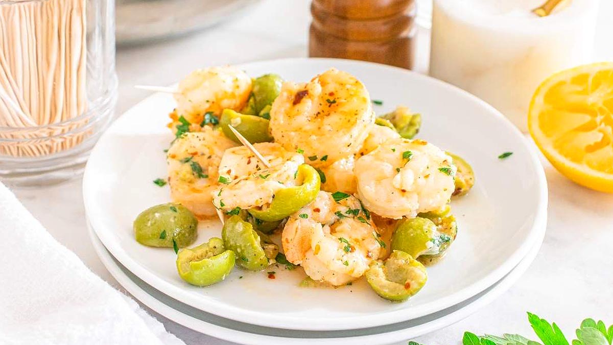 A white plate with shrimp skewers and sliced green olives, garnished with herbs, sits next to a glass, half a lemon, and a salt shaker.
