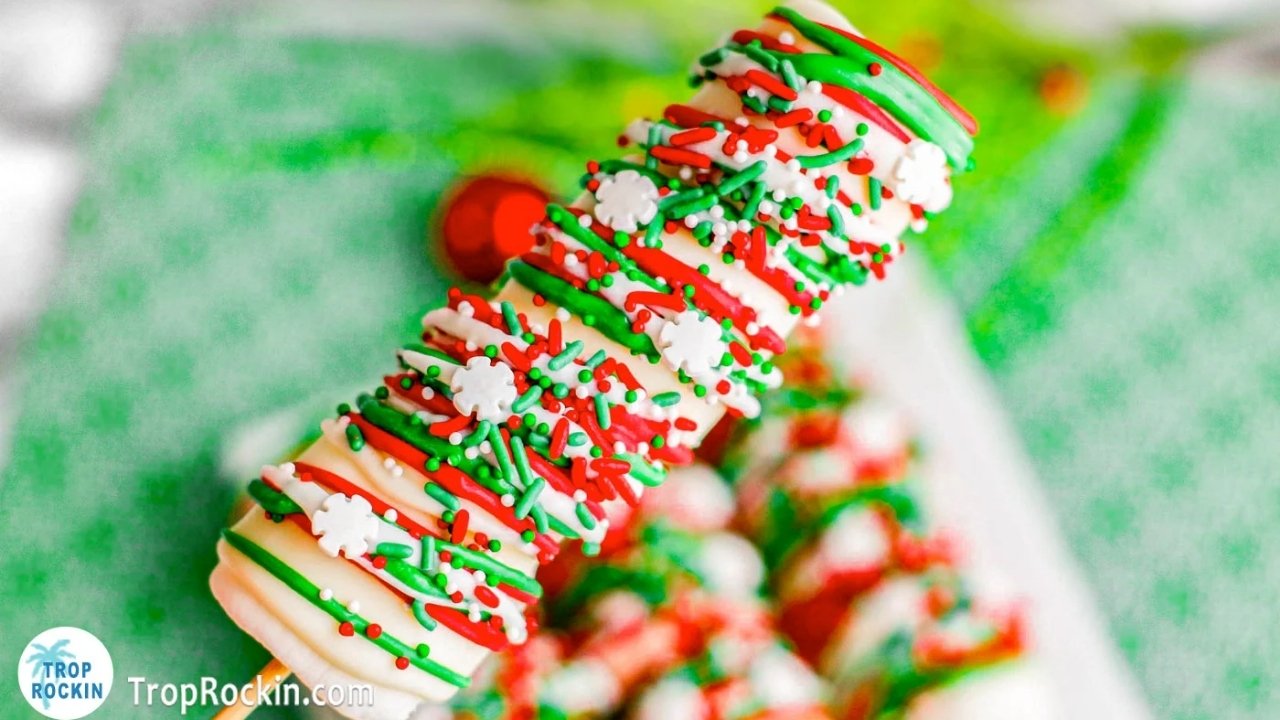 Close-up of a treat on a stick, coated in white chocolate and decorated with red, green, and white sprinkles and snowflake-shaped candies.