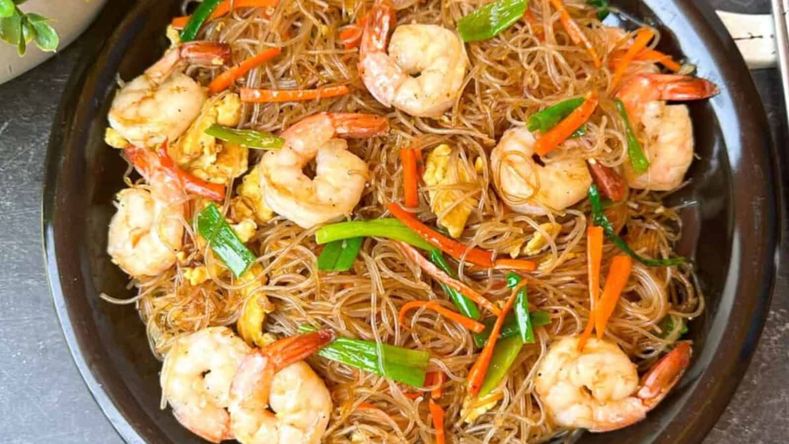 A black plate filled with stir-fried glass noodles, shrimp, sliced carrots, green onions, and scrambled egg.