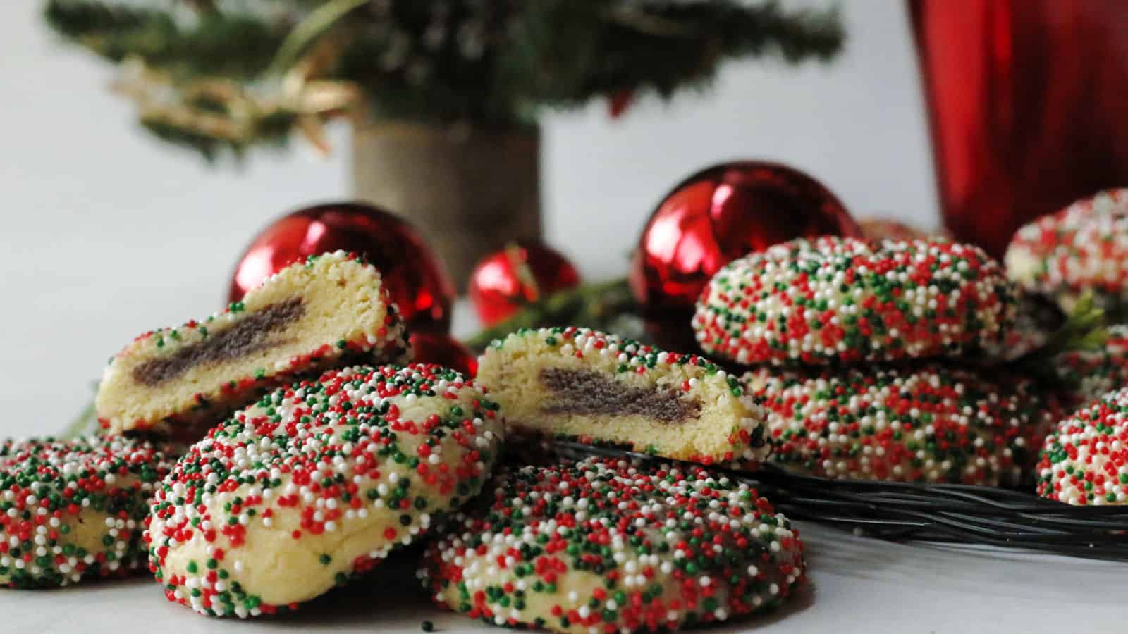 Round cookies coated with red, white, and green sprinkles are arranged on a platter, some split open to reveal a chocolate filling, with Christmas ornaments and tree in the background.