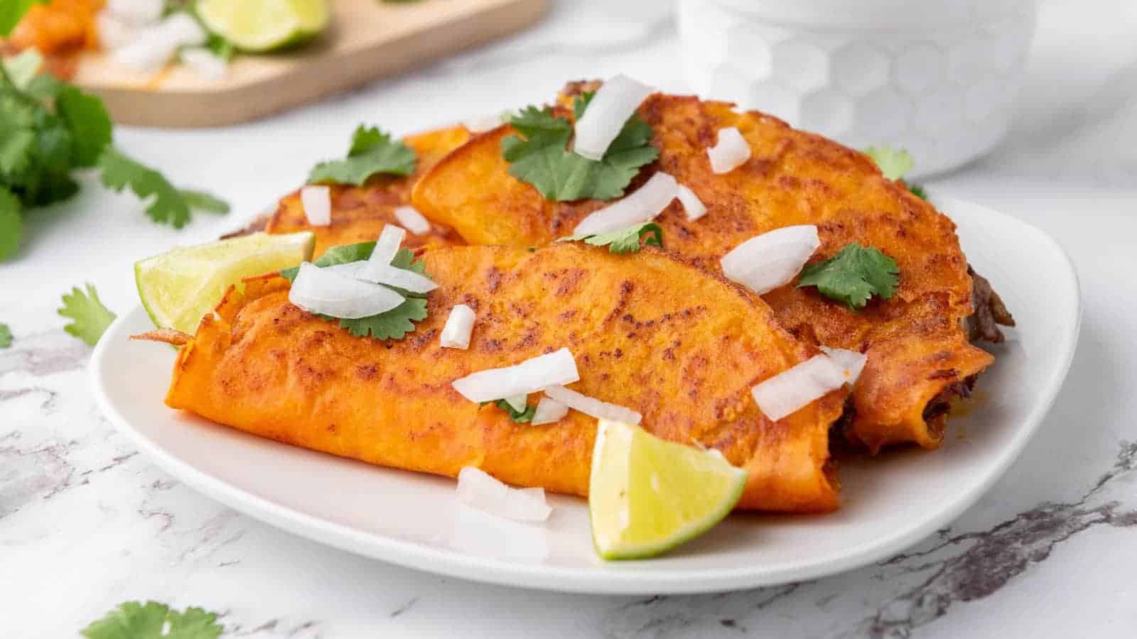 Two folded, golden-orange tacos garnished with chopped onions, cilantro, and lime wedges are served on a white plate.