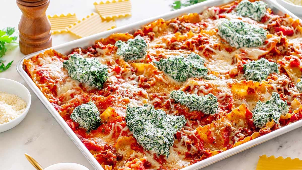 A baked lasagna in a rectangular tray, topped with dollops of spinach ricotta mixture and melted cheese, surrounded by ingredients like uncooked lasagna noodles and grated cheese.
