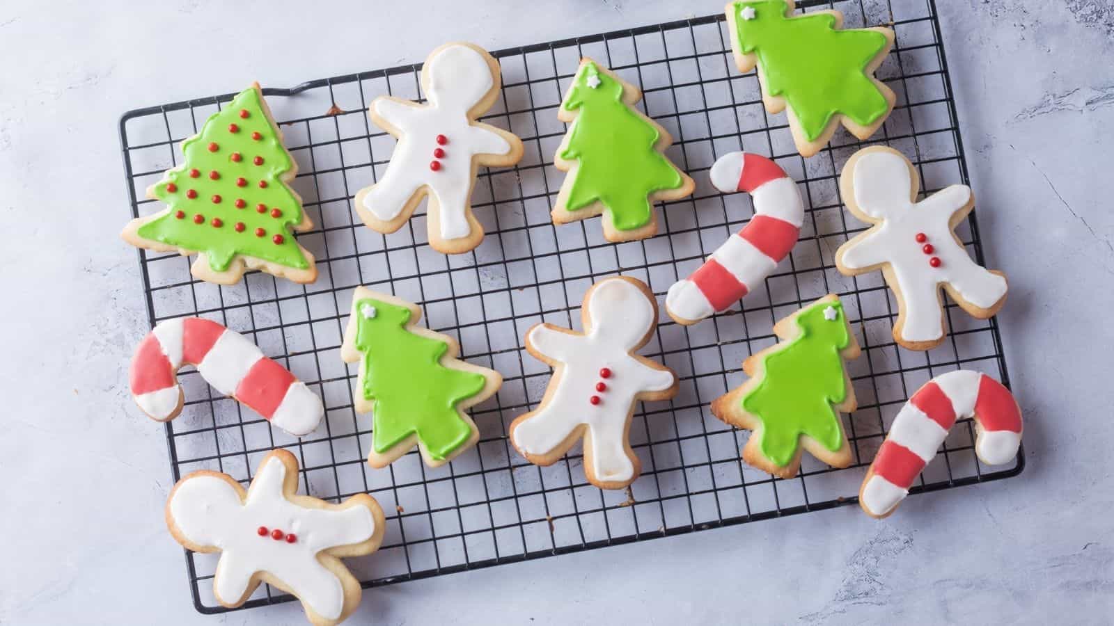 Christmas-themed sugar cookies shaped like gingerbread people, Christmas trees, and candy canes with white, green, and red icing on a cooling rack.