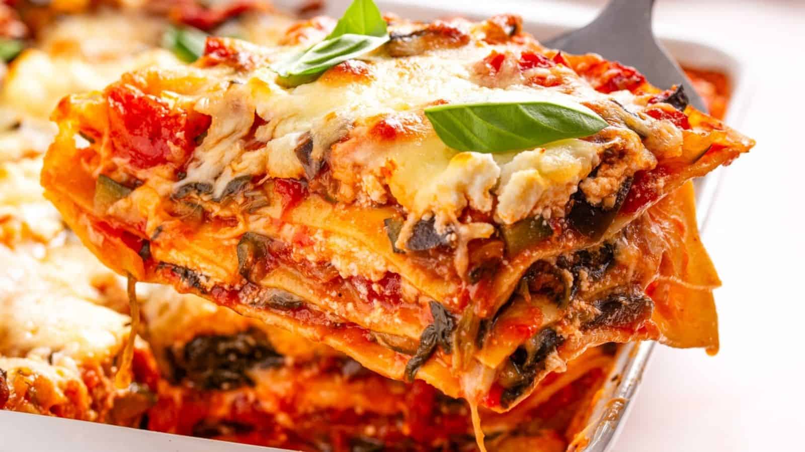A close-up of a slice of vegetable lasagna being lifted from a baking dish, topped with melted cheese and a fresh basil leaf.
