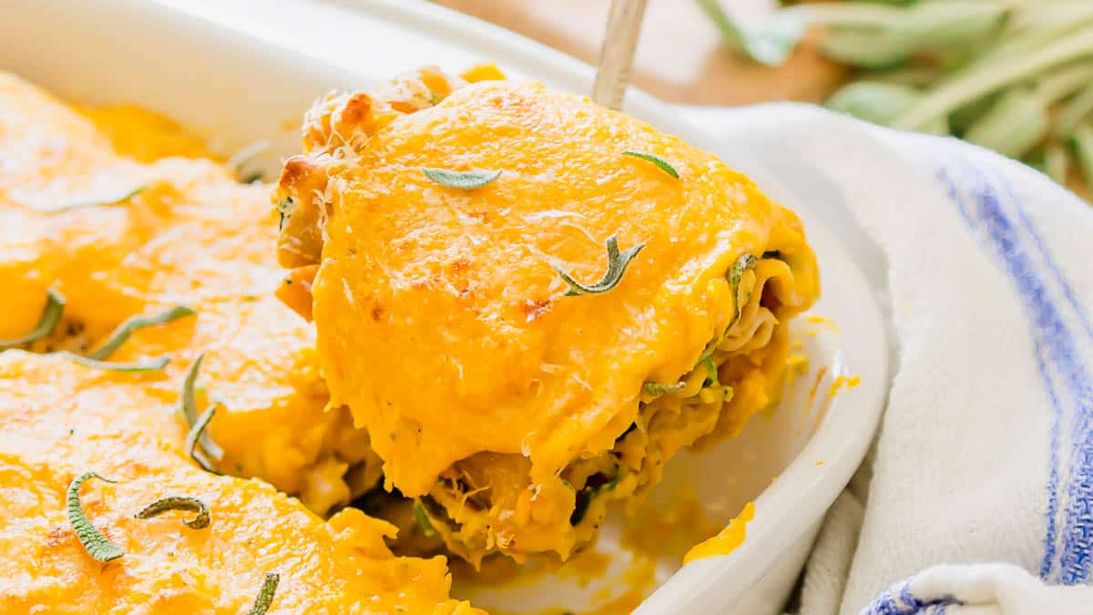 A serving of cheesy baked lasagna is being lifted from a white casserole dish, topped with melted cheese and herb garnish.