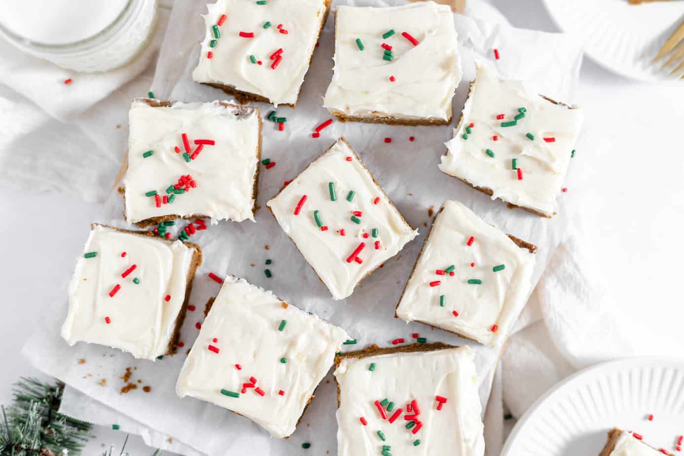 Nine frosted square cookie bars topped with red and green sprinkles are arranged on parchment paper, with plates and festive decor nearby.