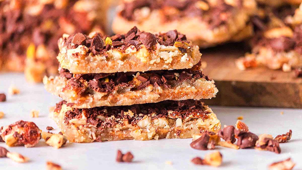 Three nut and chocolate chip dessert bars are stacked on a white surface, with crumbs scattered around and more bars visible in the background.