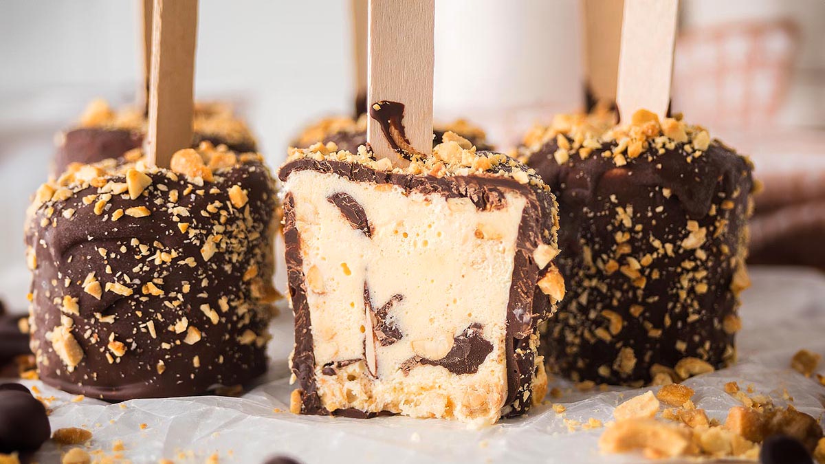 Several chocolate-covered ice cream bars with wooden sticks are coated with chopped nuts. One bar in the front has a bite taken out, revealing the inside.