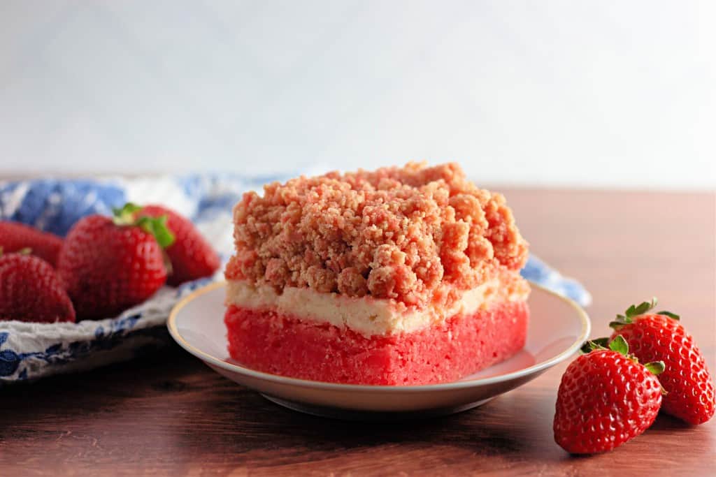 A slice of strawberry crumb cake with a crumb topping, cream layer, and pink base sits on a plate beside fresh strawberries.