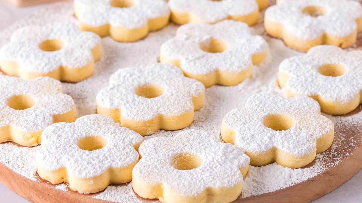 Flower-shaped cookies dusted with powdered sugar are arranged on a wooden surface with extra sugar sprinkled around them.