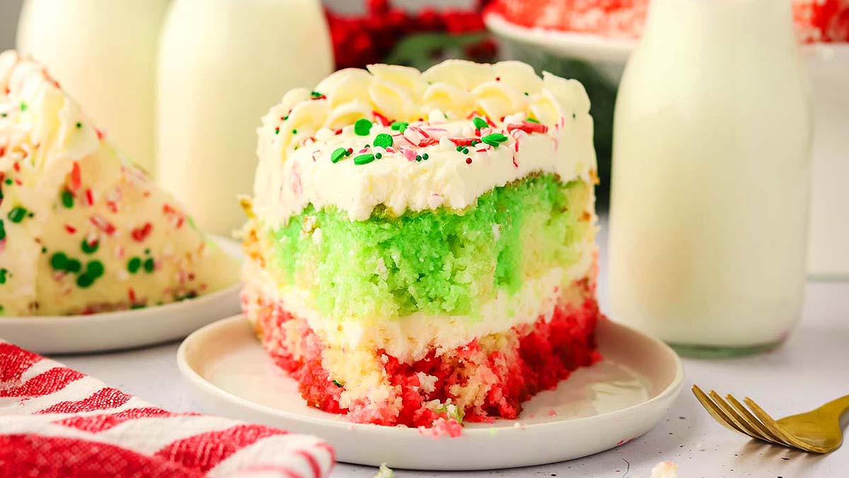 A slice of three-layer Christmas cake with red, green, and white layers, topped with white frosting and sprinkles, sits on a plate next to bottles of milk.