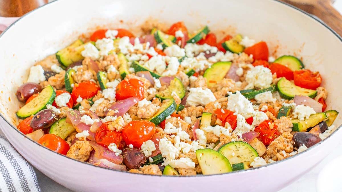 A skillet filled with cooked zucchini, cherry tomatoes, red onion, ground meat, Kalamata olives, and crumbled feta cheese.
