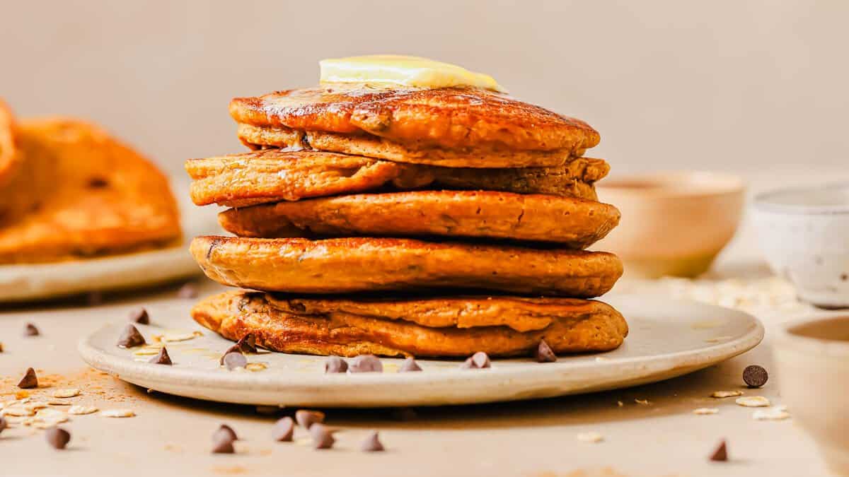 A stack of pancakes topped with a pat of butter sits on a plate, surrounded by scattered chocolate chips and oats.