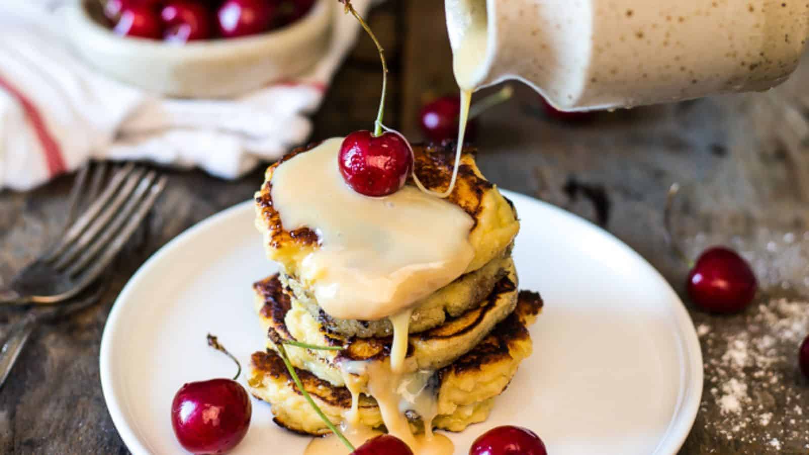 A stack of pancakes topped with cherries is drizzled with a light-colored sauce on a white plate, with cherries and cutlery nearby.