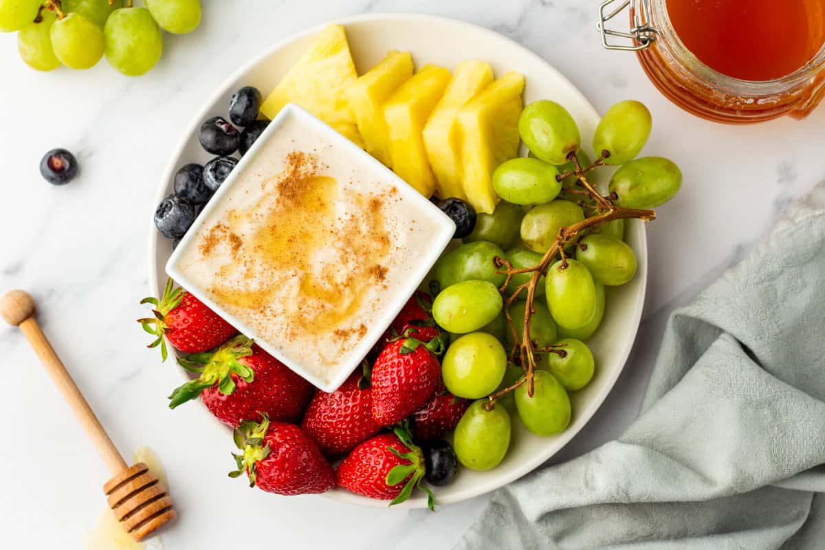 A plate with strawberries, green grapes, pineapple slices, blueberries, and a dish of yogurt topped with honey and cinnamon. A honey dipper and jar are nearby.