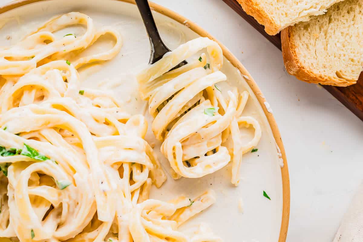 A plate of creamy fettuccine Alfredo with a fork twirling noodles, garnished with parsley, next to slices of bread on a wooden board.